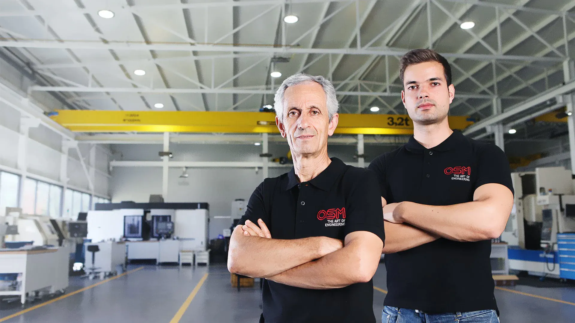 Two men in black polo shirts stand confidently with arms crossed in a spacious engineering workshop.